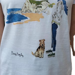 May include: White t-shirt with a watercolor illustration of a person walking a dog on a beach. The illustration includes a cliff, ocean, and the text "Elena Ciortan".