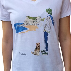 May include: White t-shirt with a watercolor illustration of a person with a dog walking on a beach with a cliff in the background. The text "Eleanor Christine" is printed on the shirt.
