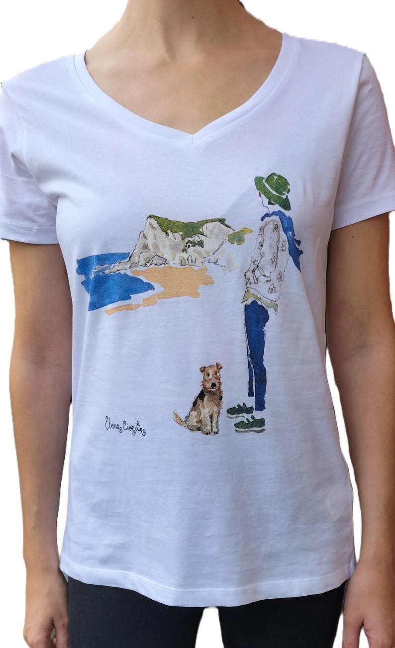 May include: White t-shirt with a watercolor illustration of a person with a dog walking on a beach with a cliff in the background. The text "Eleanor Christine" is printed on the shirt.