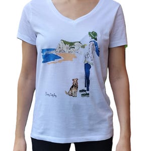 May include: White v-neck t-shirt with a watercolor illustration of a person with a dog walking on a beach with a cliff in the background. The text "Cherry  Cobbler" is printed on the bottom left of the shirt.