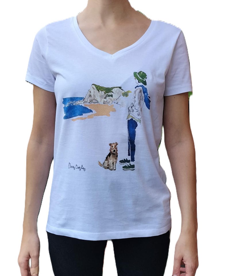 May include: White v-neck t-shirt with a watercolor illustration of a person with a dog walking on a beach with a cliff in the background. The text "Cherry  Cobbler" is printed on the bottom left of the shirt.
