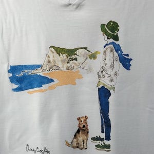 May include: White t-shirt with a watercolor illustration of a person with a dog standing on a beach. The text "Elena Ciortina" is printed on the bottom of the shirt.