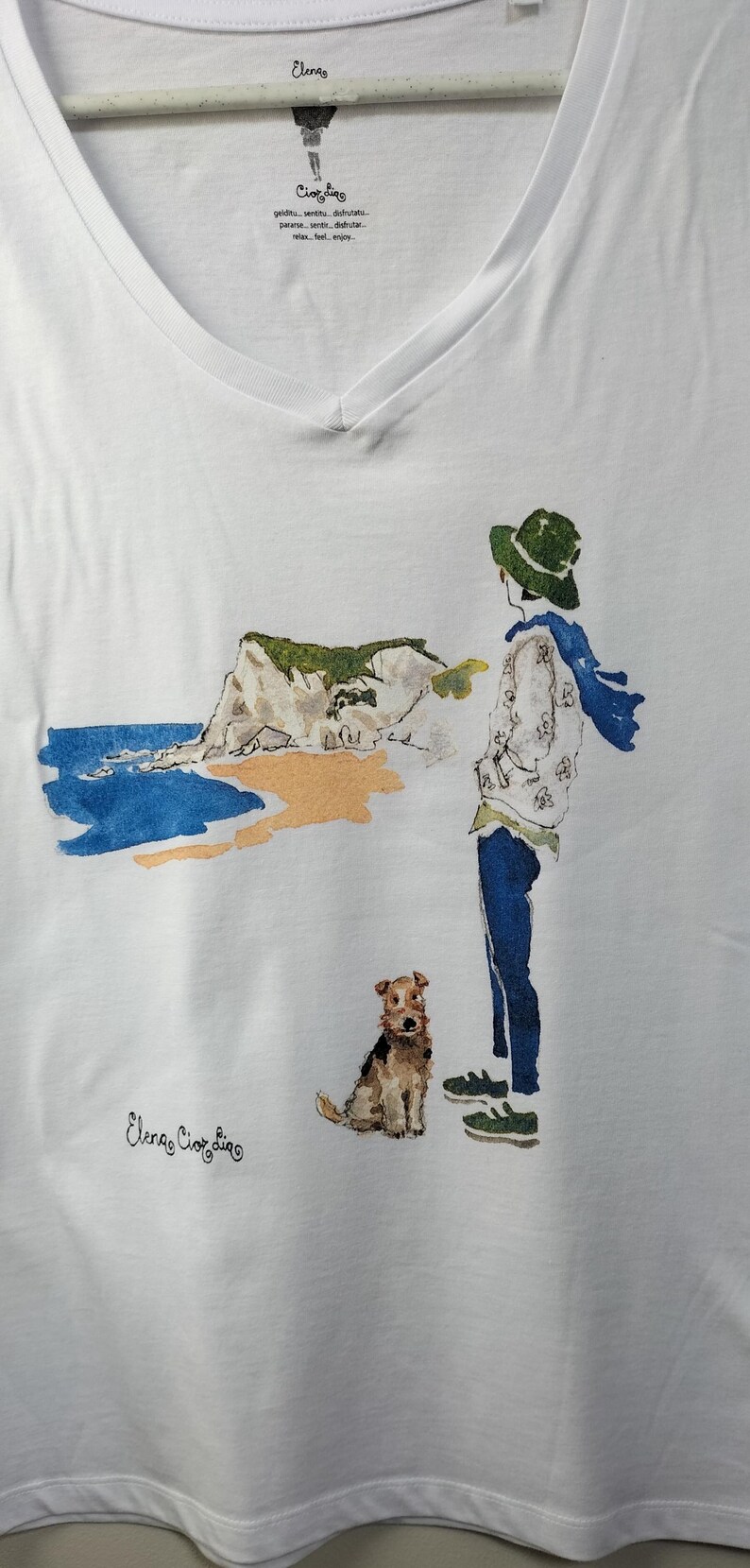 May include: White t-shirt with a watercolor illustration of a person with a dog standing on a beach. The text "Elena Ciortina" is printed on the bottom of the shirt.