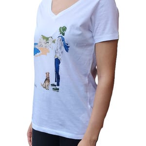 May include: White v-neck t-shirt with a graphic print of a person walking a dog on a beach. The graphic includes a white cliff, blue water, and a green hat.
