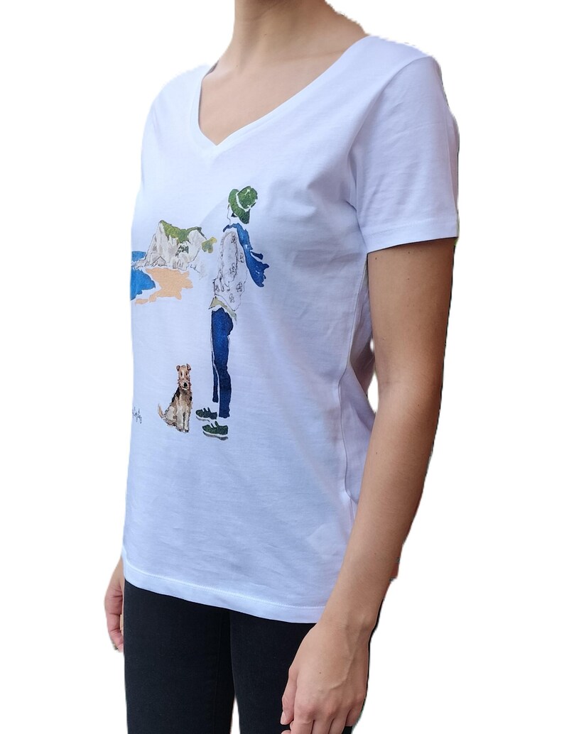 May include: White v-neck t-shirt with a graphic print of a person walking a dog on a beach. The graphic includes a white cliff, blue water, and a green hat.