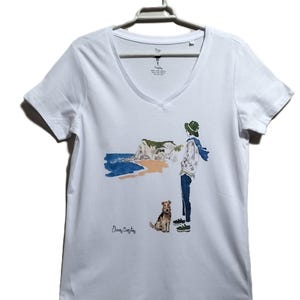 May include: White t-shirt with a watercolor illustration of a person with a dog standing on a beach with a cliff in the background. The text "Elena Copley" is printed on the shirt.
