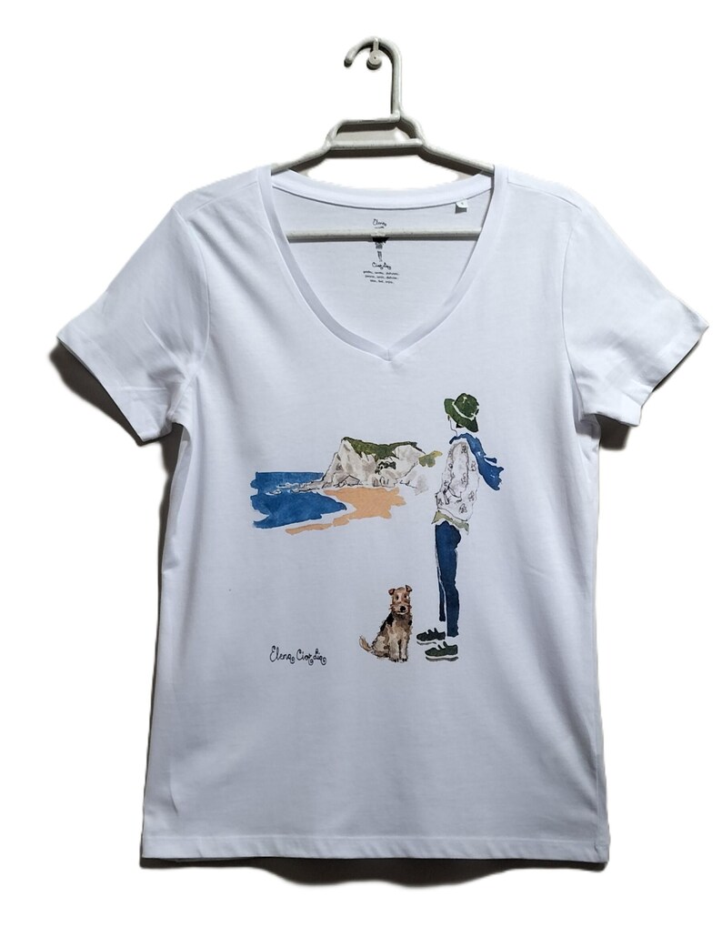 May include: White t-shirt with a watercolor illustration of a person with a dog standing on a beach with a cliff in the background. The text "Elena Copley" is printed on the shirt.