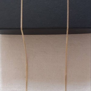 May include: A gold chain necklace with a delicate, fine link design. The chain is hanging straight down against a white fabric background.