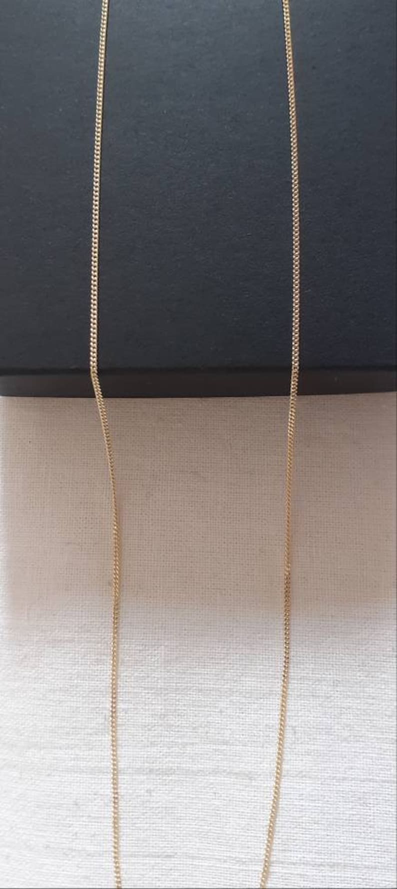 May include: A gold chain necklace with a delicate, fine link design. The chain is hanging straight down against a white fabric background.
