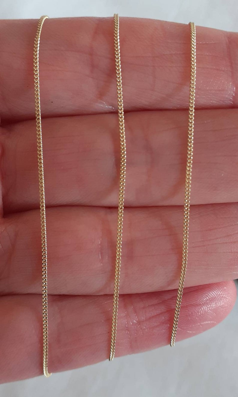 May include: Two gold chains, one on each side of the image, are shown against a light pink background. The chains are both the same length and have a delicate, fine link design.