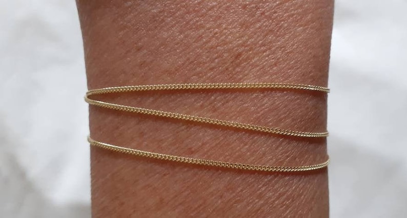 May include: A gold chain bracelet with a delicate, thin design. The bracelet is layered three times on a person's wrist.