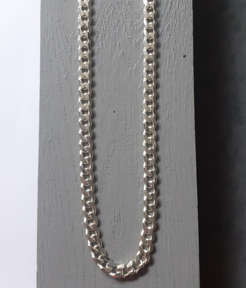 Op de afbeelding: Een zilveren curb chain ketting. De ketting is gemaakt van in elkaar grijpende schakels die een platte, rechthoekige vorm vormen.