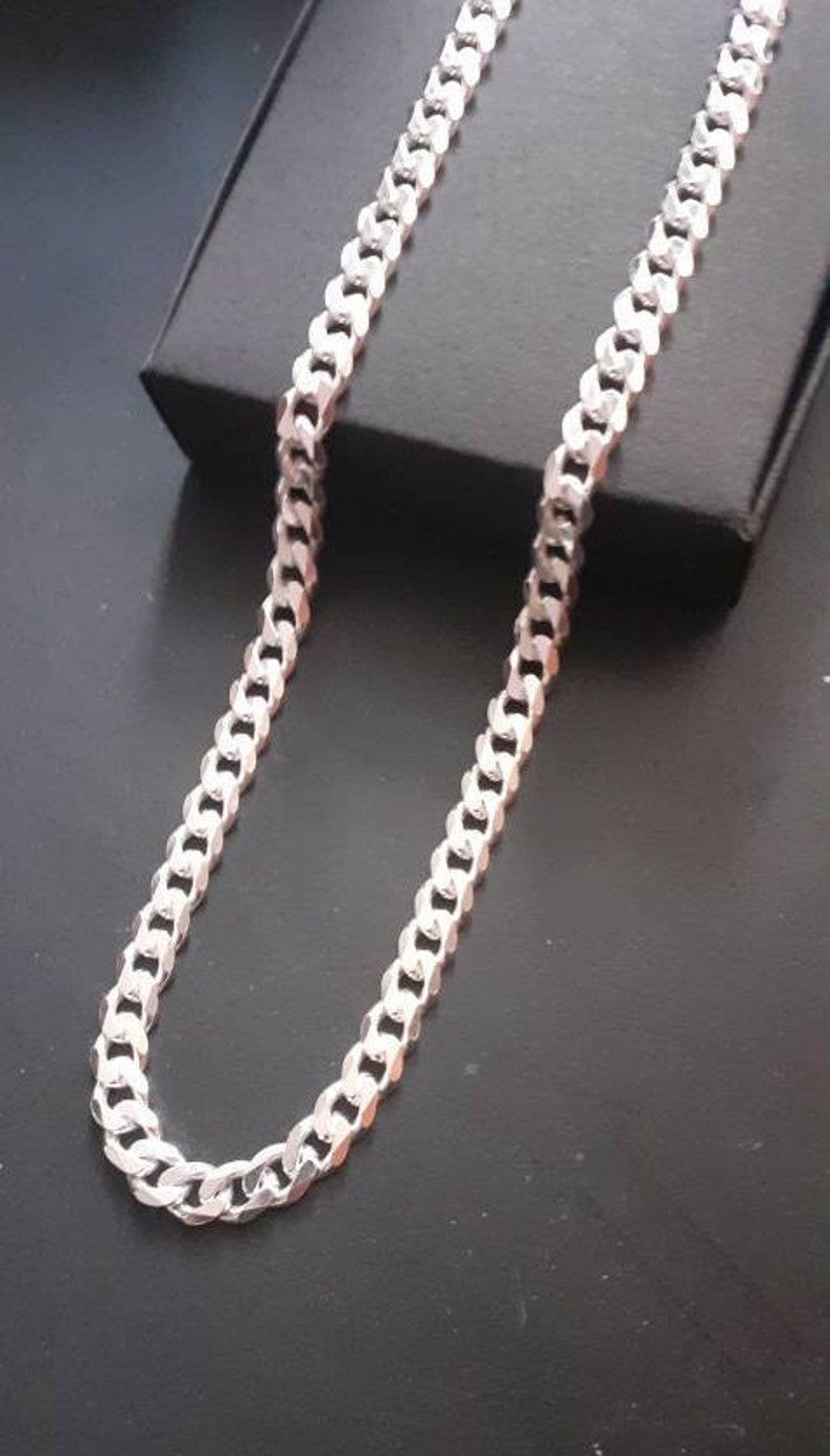 Op de afbeelding: Een zilveren curb chain ketting. De ketting is gemaakt van in elkaar grijpende schakels die een platte, rechthoekige vorm vormen.