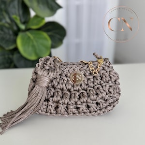 May include: A small, taupe-colored crocheted handbag with a gold chain strap and a matching tassel. The bag features a circular metal emblem with the initials "CN". The bag is sitting on a white surface.