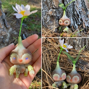 May include: Three handmade flower-themed dolls with brown and white painted faces, blue eyes, and green leaves. The dolls are made of a bulbous white material and are standing on a brown surface.