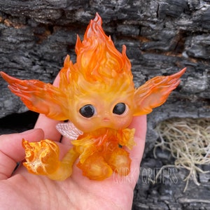 May include: A small, orange and yellow figurine of a fire elemental with wings. The figurine is made of resin and has a whimsical, cartoonish style. The figurine is sitting on a hand, with a background of a charred tree trunk.