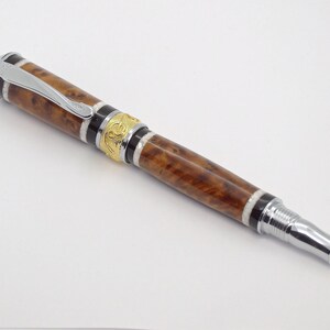 May include: A brown and gold pen with a silver clip and a silver tip. The pen has a wood grain pattern and a gold band with an intricate design.