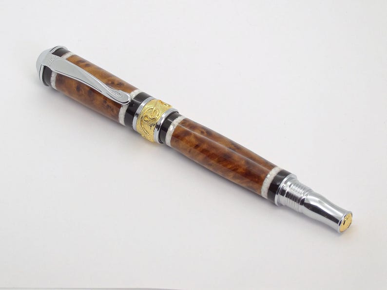 May include: A brown and gold pen with a silver clip and a silver tip. The pen has a wood grain pattern and a gold band with an intricate design.