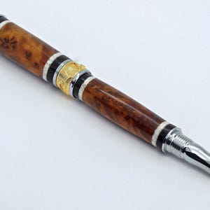 May include: A wooden pen with a brown and white burl wood design, silver accents, and a gold band. The pen has a twist mechanism.
