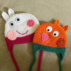 May include: Two crocheted hats with long ties. One hat is white with pink and red accents and features a cartoon pig face. The other hat is orange with green accents and features a cartoon cat face.