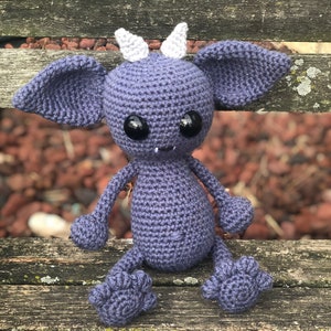 May include: A crocheted purple creature with white horns and large black eyes. The creature is sitting on a wooden bench.
