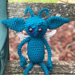 May include: A teal crocheted creature with large eyes and wings sitting on a wooden surface.