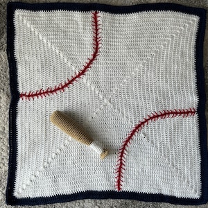 Puede incluir: Una manta de bebé de crochet blanco con un diseño de diamante de béisbol en rojo y blanco. Un pequeño bate de béisbol de crochet está encima de la manta.