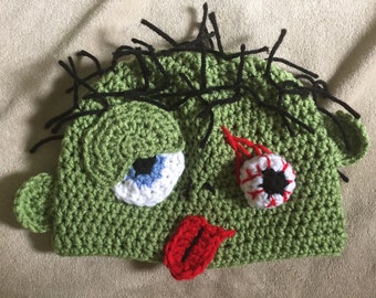 May include: Green crocheted hat shaped like a zombie face with a red tongue sticking out, one eye is white with a black pupil and the other eye is white with a black pupil and red stitching around it.