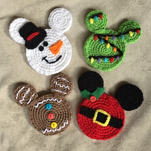 May include: Four crocheted mouse-shaped coasters with Christmas themes. The coasters are white, green, brown, and red. The white coaster has a snowman face, the green coaster has a Christmas tree with lights, the brown coaster has a gingerbread man face, and the red coaster has a Santa Claus face.
