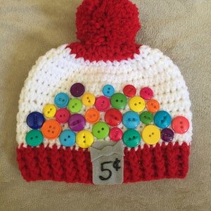 May include: White and red crocheted hat with a red pom-pom on top. The hat is decorated with colorful buttons that resemble a gumball machine. The hat has a gray felt tag with the text "5¢" on it.