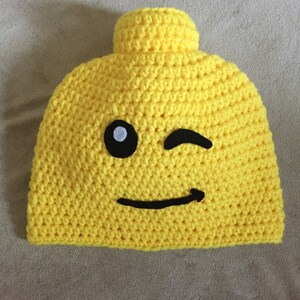 May include: Yellow crocheted hat shaped like a Lego brick with a black winking eye and a black smiling mouth.