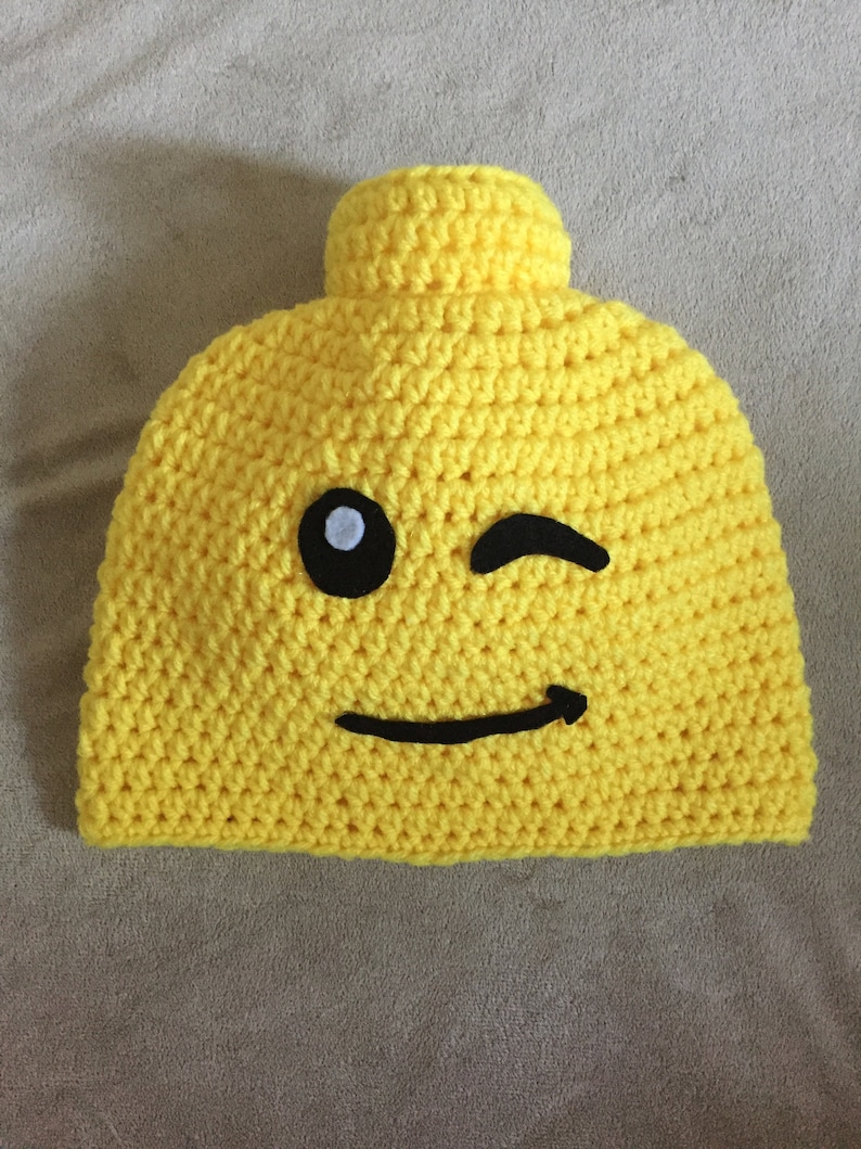 May include: Yellow crocheted hat shaped like a Lego brick with a black winking eye and a black smiling mouth.