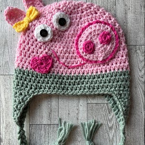 May include: A handmade crochet hat in the shape of a pig. The hat is primarily pink with a green band at the bottom and green braided ties. It features white button eyes, a pink snout, and a yellow bow.