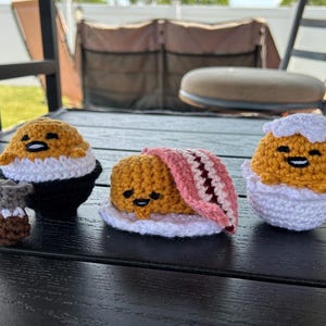 May include: Three crocheted egg-themed figures. One is wearing a black suit with a coffee cup, another is covered with a pink blanket, and the third is in an eggshell. Each has a yellow yolk and white accents.