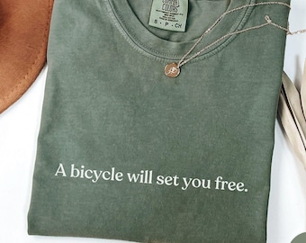 May include: A moss green crew neck t-shirt with the white text A bicycle will set you free printed on the front. The shirt features a Comfort Colors brand label at the inner neckline and is paired with a thin gold chain necklace.