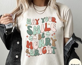 May include: A beige t-shirt with a retro-style graphic that says "May your smile be merry & bright". The graphic features colorful letters and ornaments.