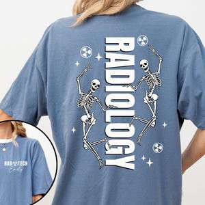 May include: Blue t-shirt with the word "RADIOLOGY" in white vertical text. Two dancing skeleton figures are on the shirt, along with radiation symbols and stars. The shirt also has the text "RAD TECH Emily" on the front.