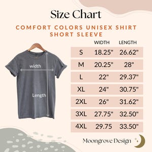 May include: A size chart for a Comfort Colors unisex short sleeve shirt. The chart lists sizes S through 4XL with width and length measurements in inches. The shirt is gray and hangs on a wooden hanger.