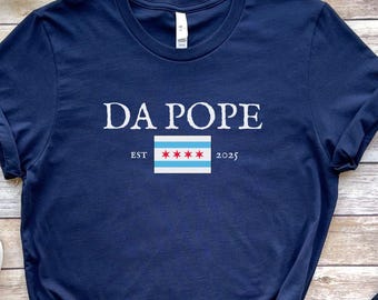 Könnte beinhalten: Marineblaues T-Shirt mit dem weißen Schriftzug "DA POPE" über einem Chicago-Flaggen-Design. Das Shirt enthält auch "EST" und "2025" in weißer Schrift. Das T-Shirt besteht aus einem weichen Material.