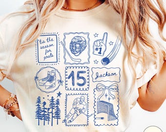 May include: An ivory t-shirt featuring a blue graphic print with a grid of hockey-themed illustrations including a helmet, a foam finger with #1, a hockey puck, skates, the number 15, a hockey player, a net, and the text 'tis the season for goals' and 'Jackson'.