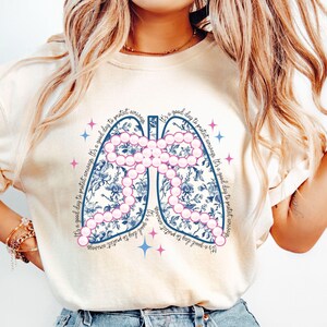 May include: Cream-colored sweatshirt featuring a stylized lung design filled with blue floral patterns and pink bead-like accents. The phrase "It's a good day to protect airways" is printed around the lung graphic. The design includes blue and pink star accents.