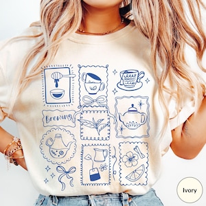 May include: Ivory-colored sweatshirt with a blue tea-themed graphic. The design features various tea-related illustrations, including teacups, teapots, honey, and tea leaves, arranged in a grid pattern. The word "brewing" is also included in the design.