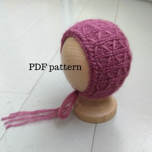 Peut inclure: Un bonnet de bébé tricoté de couleur rose poudré, orné d'un motif géométrique. Le bonnet est présenté sur une tête en bois et possède des liens. Le texte "PDF pattern" est visible.