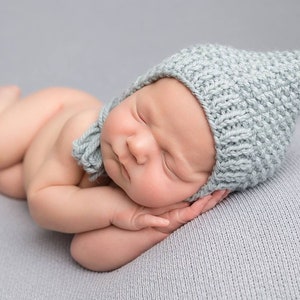 May include: A newborn baby wearing a light blue knitted hat with a pointed tip, sleeping on a light blue blanket. The baby is curled up with their hands under their chin.