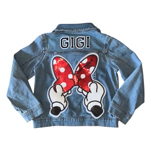 May include: Light blue denim jacket with a sequined Minnie Mouse bow and hands design on the back. The name "GIGI" is embroidered above the bow in black and white lettering. A classic, stylish jacket.
