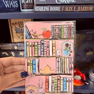 May include: A pink bookmark with a colourful bookshelf illustration, including a lamp, apple, rose, crown, and art supplies. The bookmark is held in a hand, with books visible in the background.