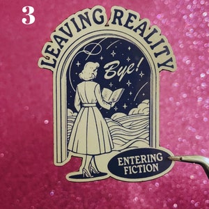 May include: A sticker with a retro illustration of a woman walking through a doorway labeled "Leaving Reality" and entering a doorway labeled "Entering Fiction". The woman is holding a book and looking up at the stars. The sticker is on a pink background.