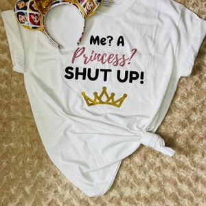 May include: White t-shirt with the text "Me? A Princess! Shut Up!" and a gold crown graphic. A pair of gold mouse ears with a collage of Disney princesses is on top of the shirt.