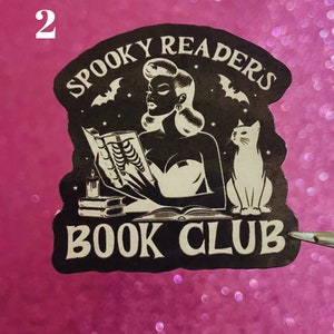May include: Black and white sticker with a woman reading a book, a cat, and bats. The text reads "Spooky Readers Book Club".