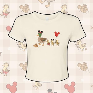 May include: Cream-coloured t-shirt featuring a cartoon duck family wearing Mickey Mouse ears and holding a red balloon. The ducklings are wearing red shorts. The shirt has a crew neck and short sleeves. The background has a pattern of ducks, Mickey Mouse cookies, and red balloons.